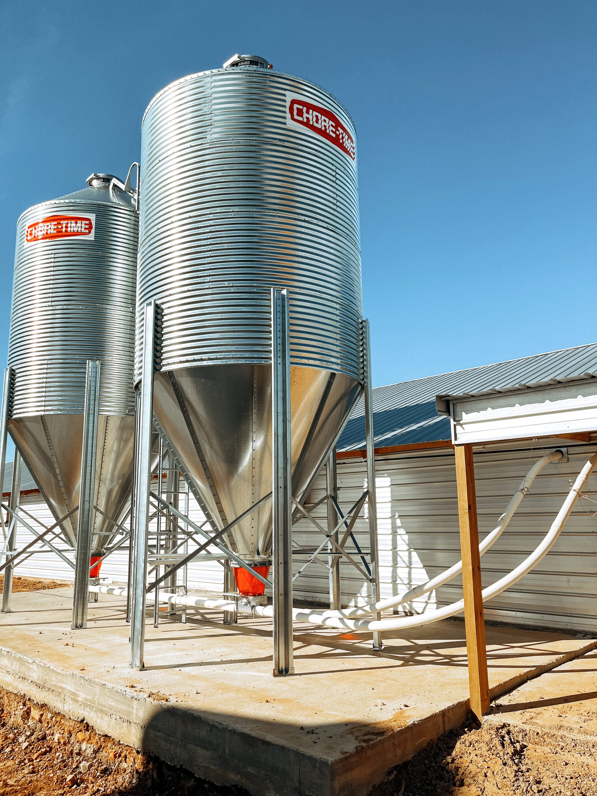 Two galvanized Chore-Time feed bins on a concrete pad beside a poultry house
